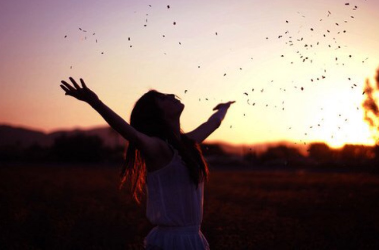 Une femme aux bras tendus dans un champ au coucher du soleil, exprimant sa spiritualité.
