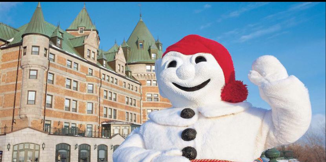 Une mascotte de bonhomme de neige du Carnaval de Québec 2019 agite devant un grand bâtiment.