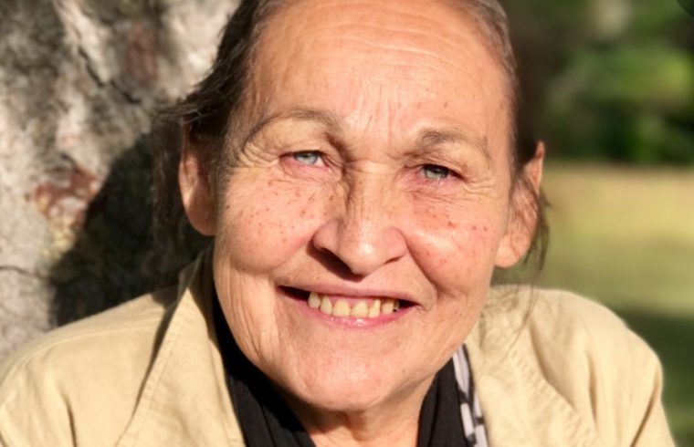 Une femme plus âgée sourit en s’appuyant contre un arbre à Uiesh, capturant l’essence de Quelque part de Joséphine Bacon.