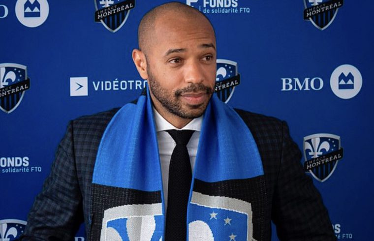 Un homme en costume et écharpe bleue ressemblant à Thierry Henry debout devant un fond bleu.