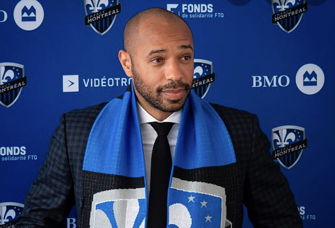 Un homme en costume et écharpe bleue ressemblant à Thierry Henry debout devant un fond bleu.