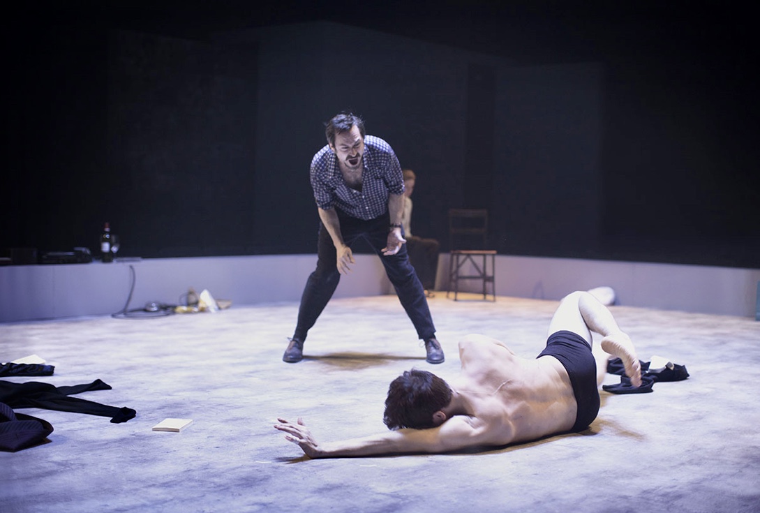 Un homme joue une scène au sol tandis qu'un autre homme se tient à côté de lui dans un décor théâtral unique.