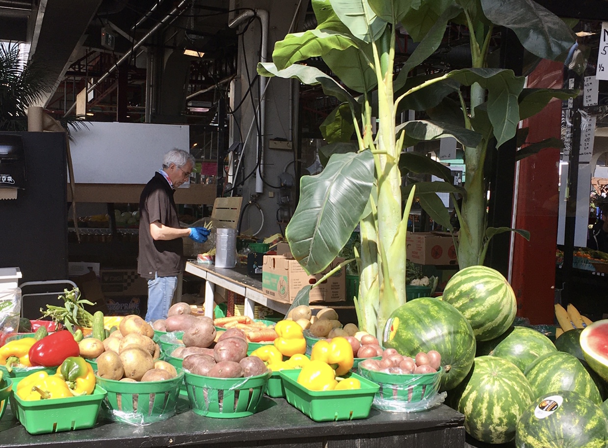Un homme debout devant un étalage de fruits et légumes, mettant en valeur l’abondance de produits frais dans le contexte de la pandémie de COVID-19 et du retour progressif à la vie à Montréal.