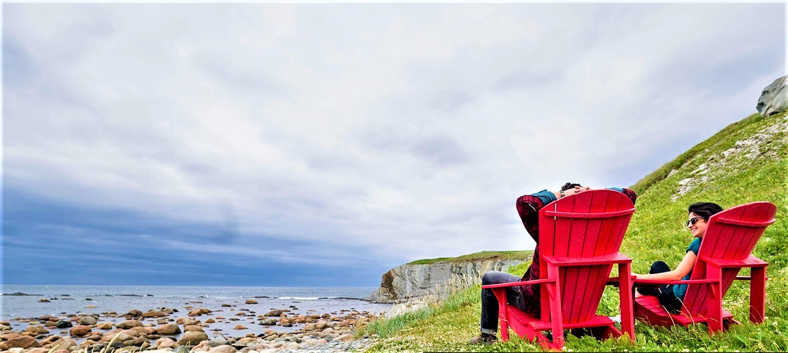 Deux personnes, profitant du tourisme au Canada, assises sur des chaises Adirondack rouges sur une colline herbeuse.