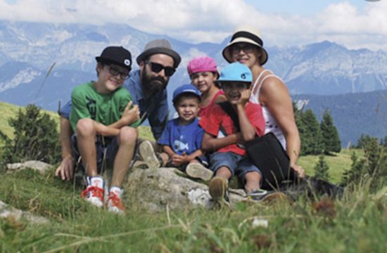 Une famille pose sur une colline herbeuse avec des montagnes en arrière-plan, capturant leurs voyages aventureux.