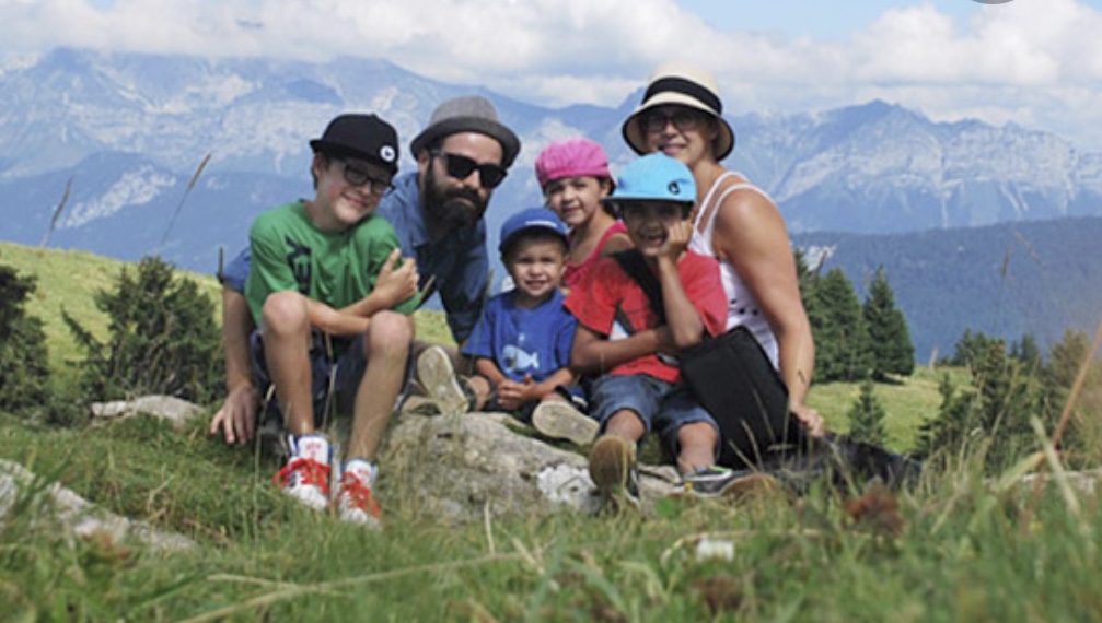 Une famille pose sur une colline herbeuse avec des montagnes en arrière-plan, capturant leurs voyages aventureux.