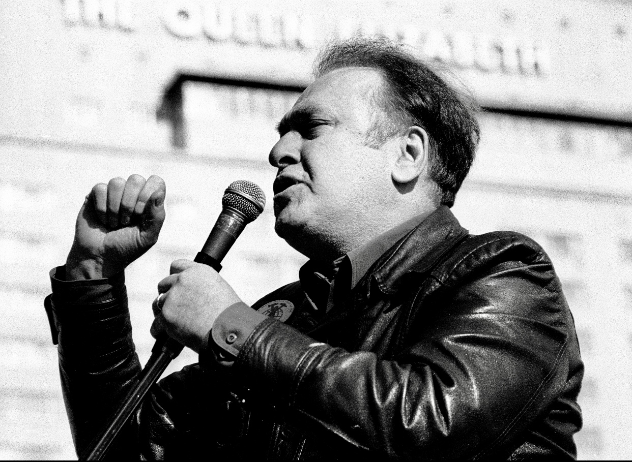 Un homme vêtu d'une veste en cuir parlant dans un microphone tout en étant photographié.
