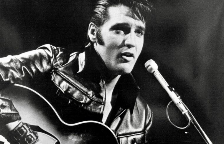 Elvis Presley, le musicien légendaire, dégage sans effort son style emblématique en portant une veste en cuir noire tout en jouant sur une guitare acoustique.
