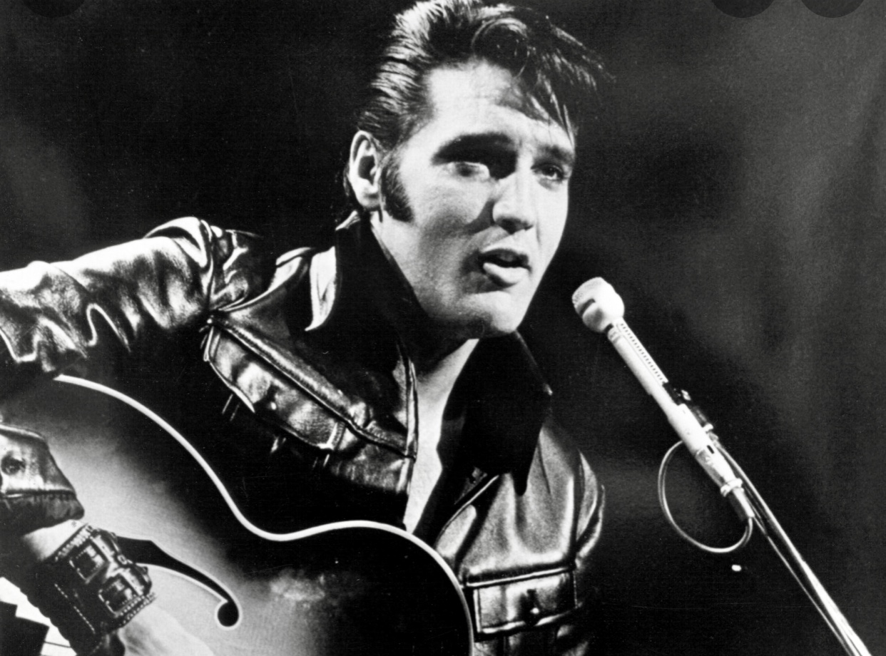 Elvis Presley, le musicien légendaire, dégage sans effort son style emblématique en portant une veste en cuir noire tout en jouant sur une guitare acoustique.