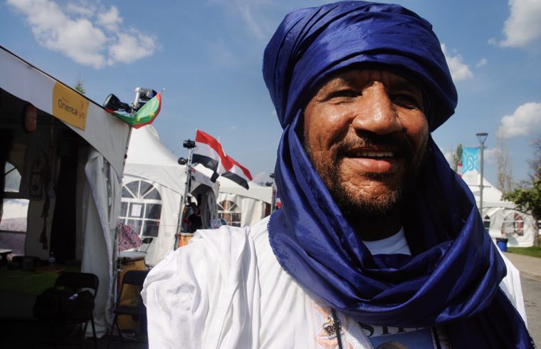 Un homme au turban bleu sourit au festival Orientalys, entouré de tentes.