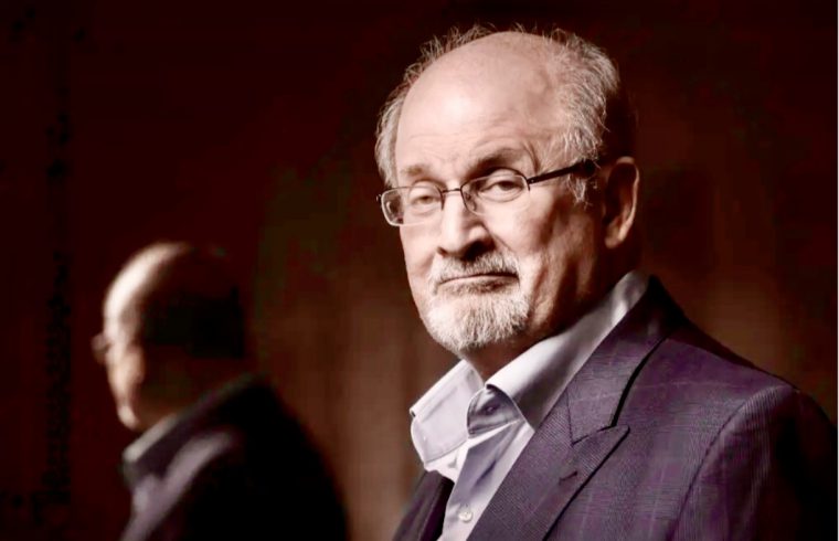 Un homme en costume et lunettes, ressemblant à Salman Rushdie, se tient devant un miroir.