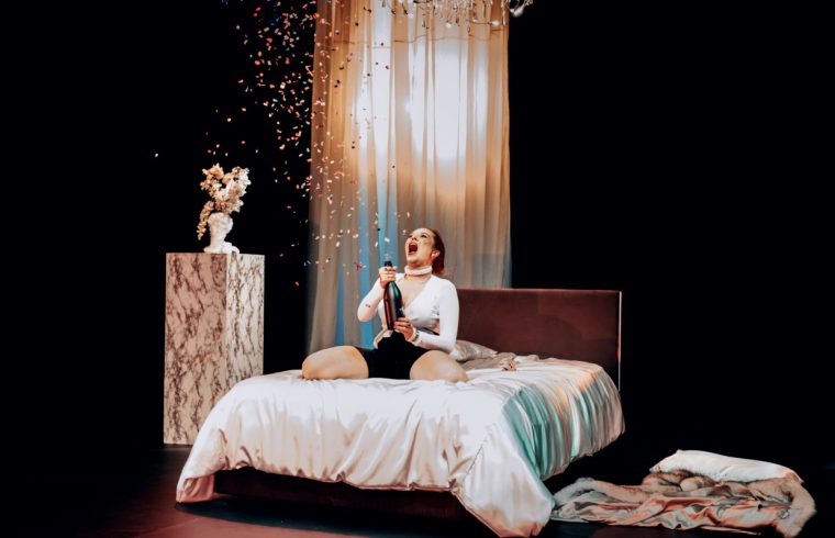 Une femme assise sur un lit avec des confettis tombant du plafond, créant une atmosphère fantaisiste de théâtre.