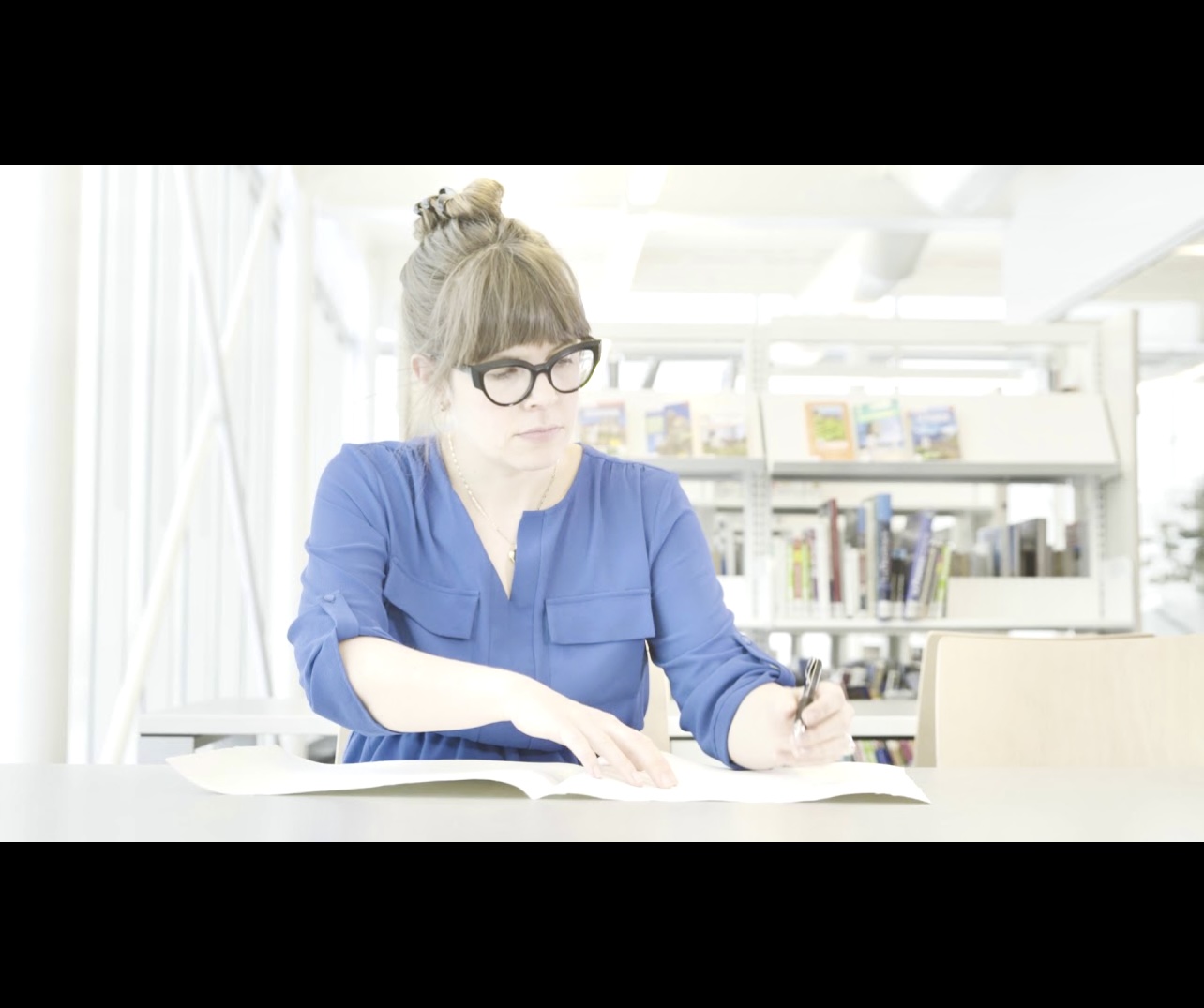 Une femme à lunettes écrivant un livre sur la Littérature québécoise dans une bibliothèque.
