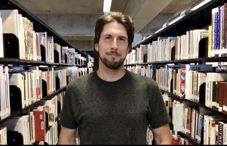 Un homme debout devant une étagère remplie de Littérature québécoise dans une bibliothèque.