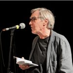 Un auteur, nominé pour le Prix de la poésie 2023, tient passionnément un livre tout en prononçant un discours captivant devant un micro.
