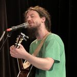 Un homme jouant de la guitare acoustique en chantant dans un micro lors du Prix de la poésie 2023.