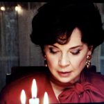 Anne Dorval, actrice connue pour ses performances captivantes, est élégamment vêtue d'une superbe robe rouge alors qu'elle allume délicatement des bougies devant une télévision.