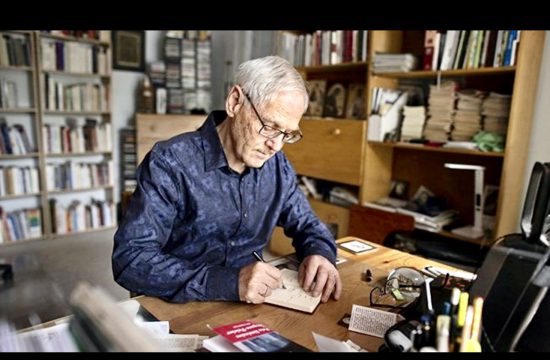 Jacques Gauthier, un homme plus âgé, assis à un bureau avec un cahier, compose son chef-d'œuvre "Le refrain des heures.