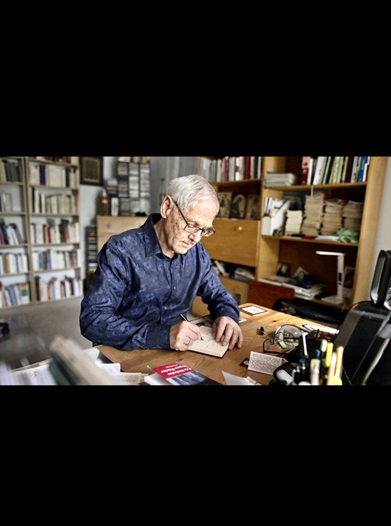 Jacques Gauthier, un homme plus âgé, assis à un bureau avec un cahier, compose son chef-d'œuvre "Le refrain des heures.