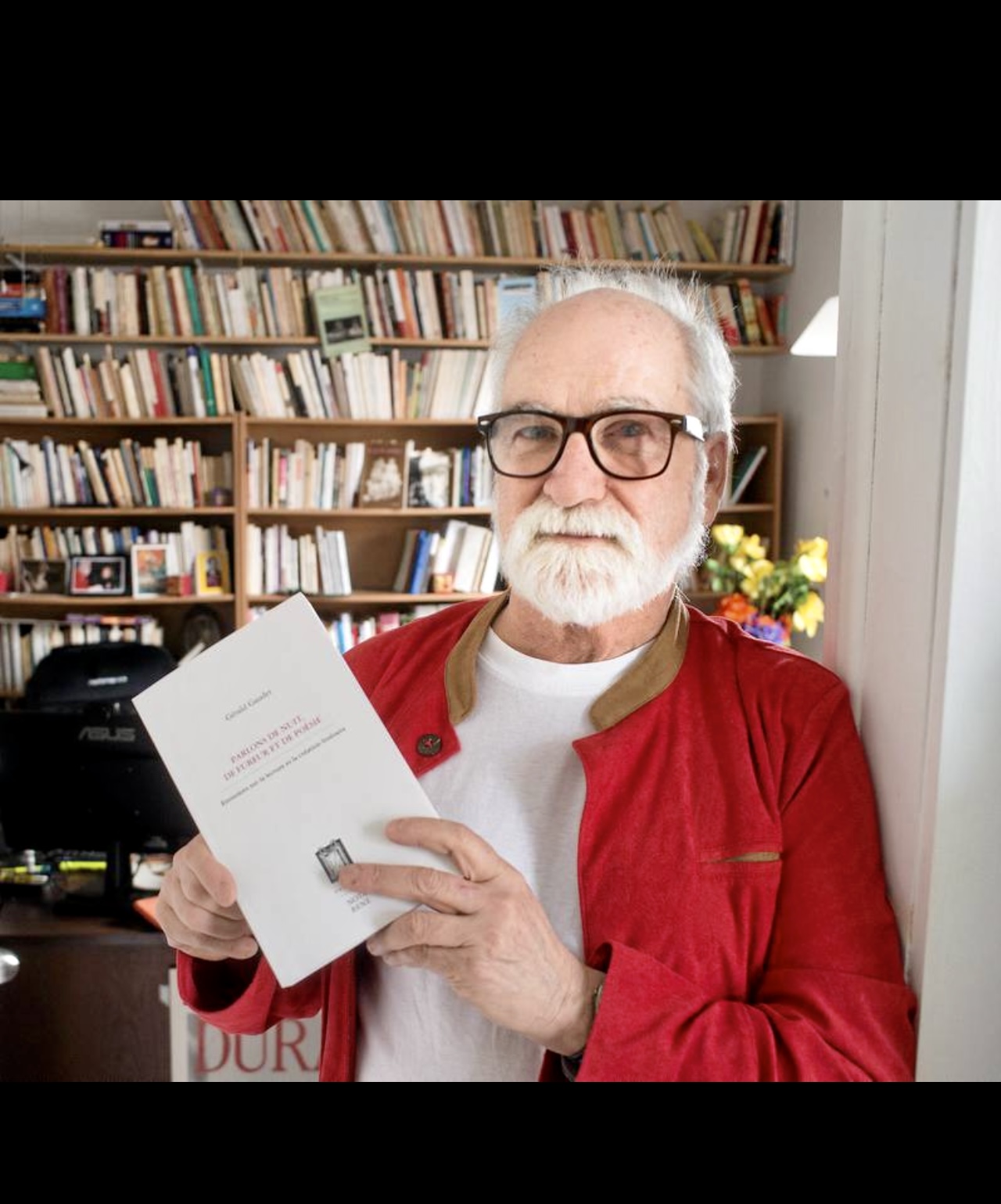 Un homme plus âgé tenant un livre devant une étagère lors d'une entrevue avec Gérald Gaudet.